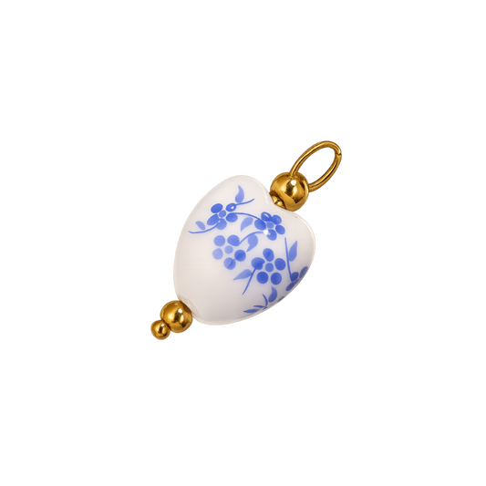 A heart-shaped white ceramic pendant charm decorated with blue floral and berry motifs, finished with polished gold-tone metal caps and a top loop.