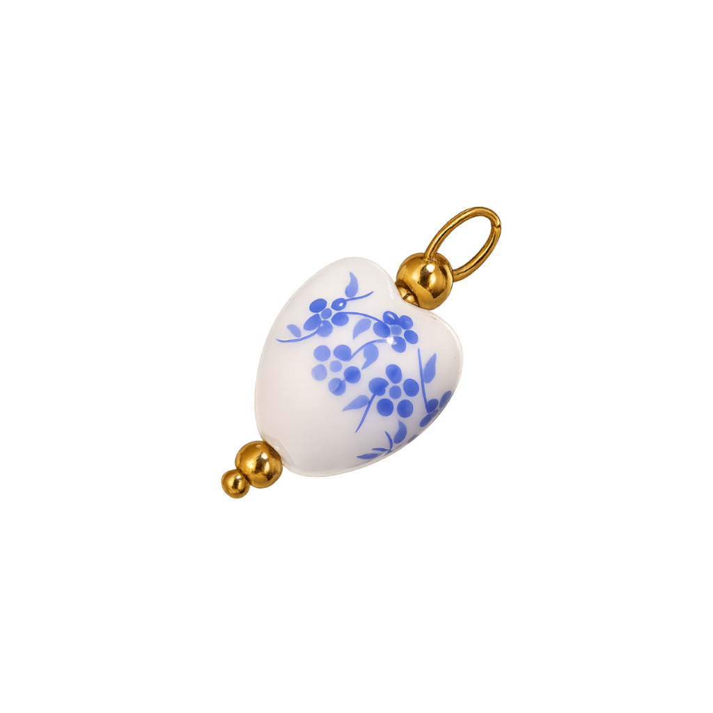 A heart-shaped white ceramic pendant charm decorated with blue floral and berry motifs, finished with polished gold-tone metal caps and a top loop.