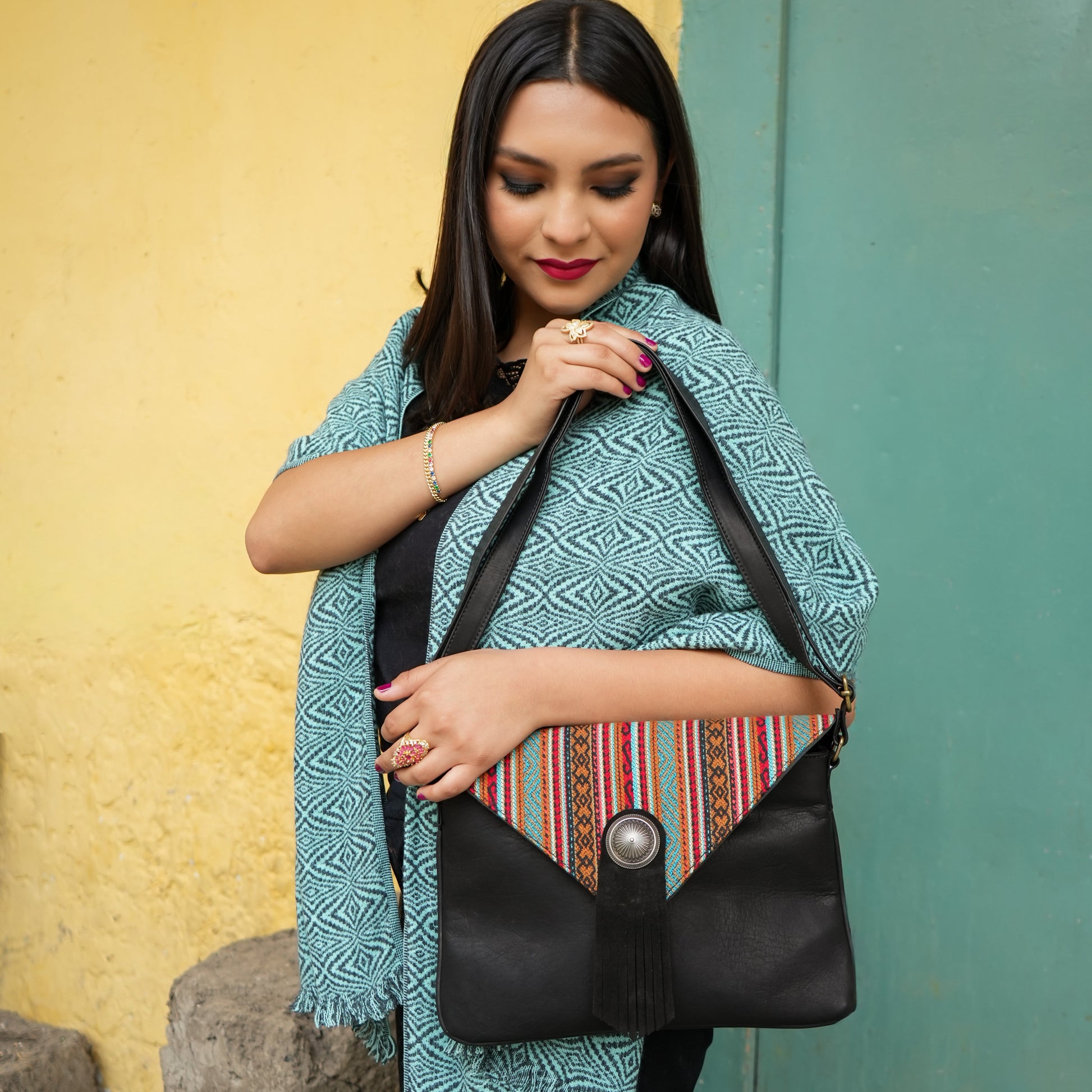 Black shoulder bag with a colorful woven pattern and a silver-tone button closure, set against a split yellow and teal wall background.