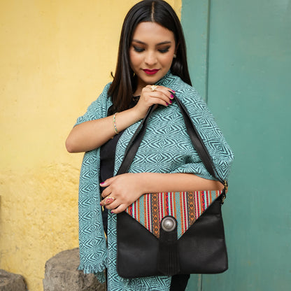 Black shoulder bag with a colorful woven pattern and a silver-tone button closure, set against a split yellow and teal wall background.