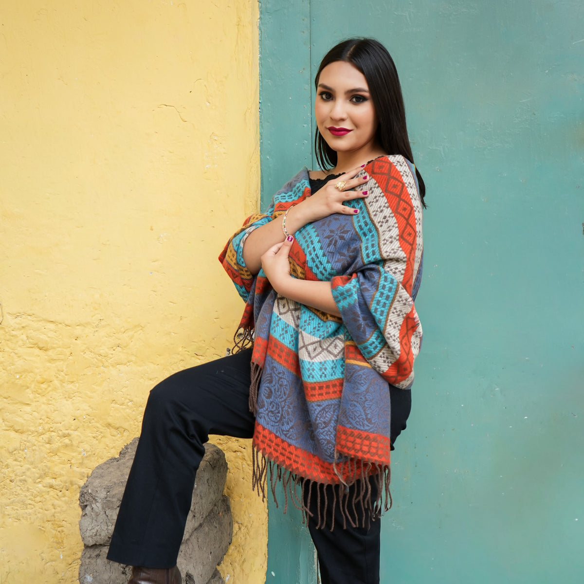 Colorful patterned knit poncho with fringe, paired with black pants and brown ankle boots, standing against a split yellow and teal wall.